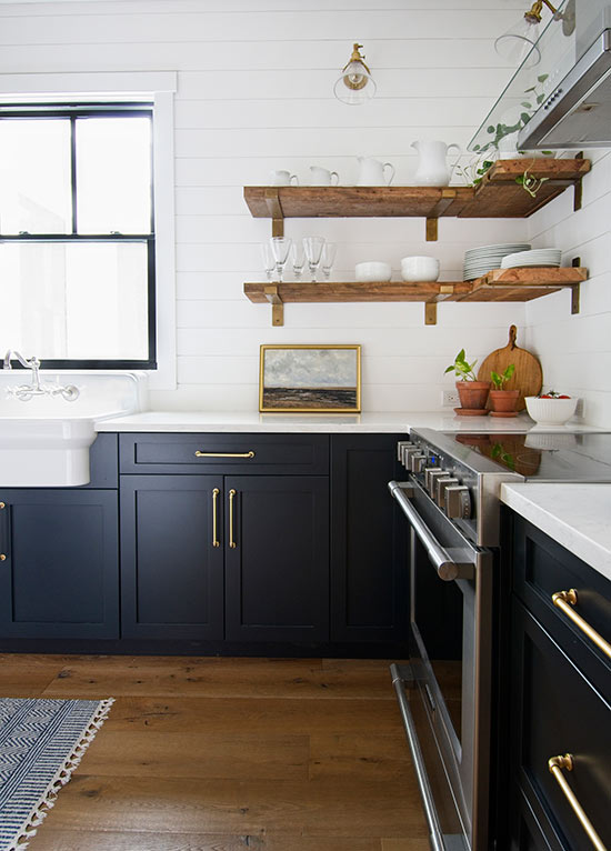 modern farmhouse kitchen with dark navy blue cabinets with gold brass hardware and open shelving with shiplap backsplash and farmhouse apron sink in front of black window