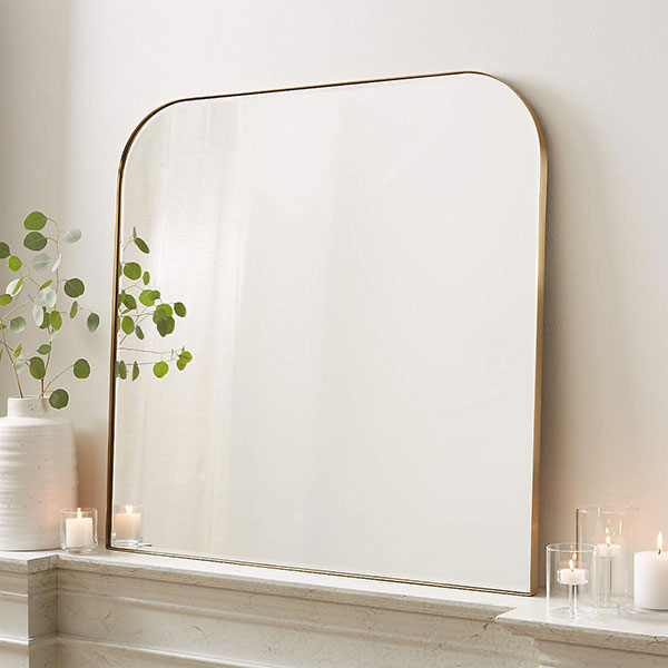 gold brass arched wall mirror leaning against white wall on fireplace mantel next to votive candles and a white vase with dried florals