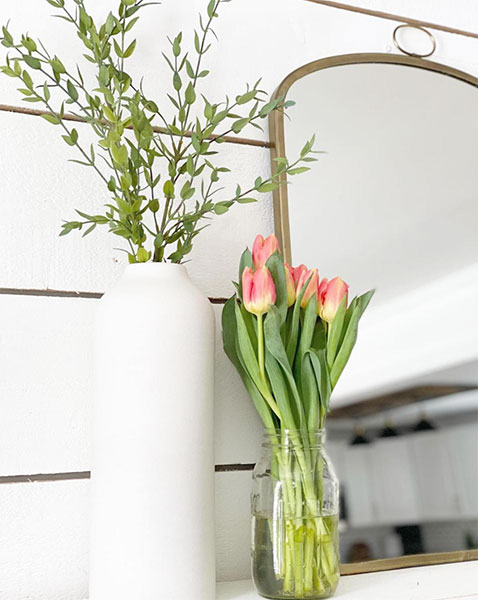 brass arched mirror on top of dresser in front of white shiplap wall next to white vase with flowers