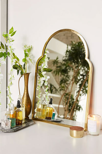 gold brass arched mirror leaning against white wall on top of dresser next to vases with dry florals
