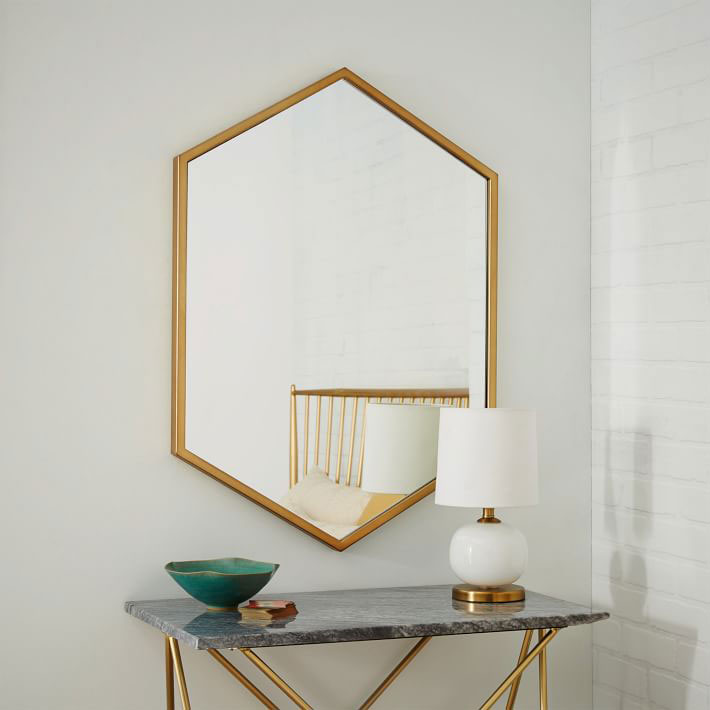 gold brass hexagon mirror on white wall above glass console table with small lamp